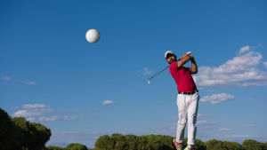 A golfer in a red shirt and white pants swings a golf club, with a golf ball in mid-air against a clear blue sky and a backdrop of trees.